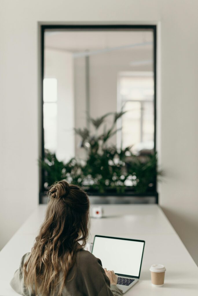 A woman with curly hair works on a laptop, facing a window with plants, symbolizing remote work.